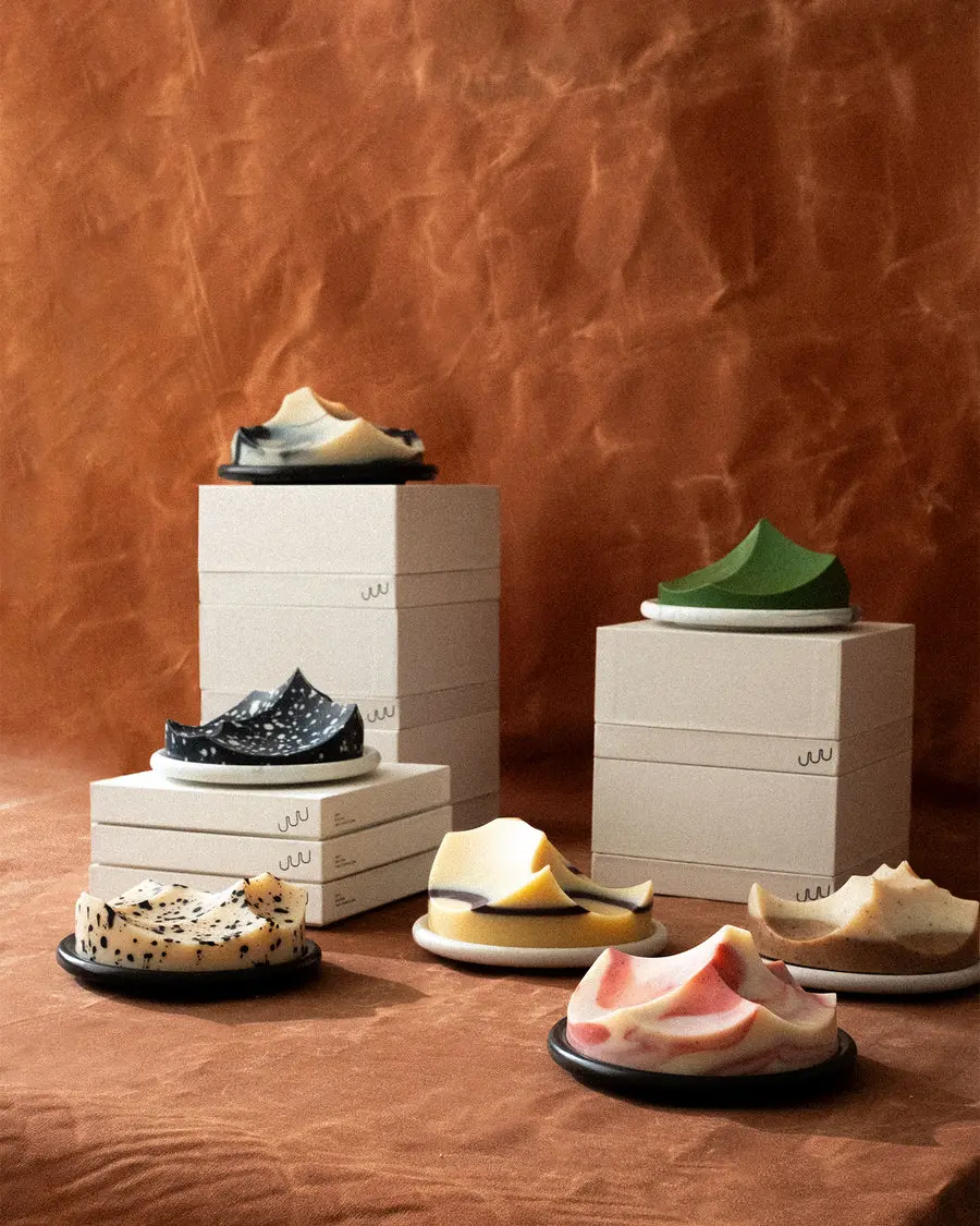 Assorted UMÉ hand-cast Erode soaps in various colors displayed on round bouton plates, arranged with stacked beige UMÉ boxes against a warm brown textured backdrop.