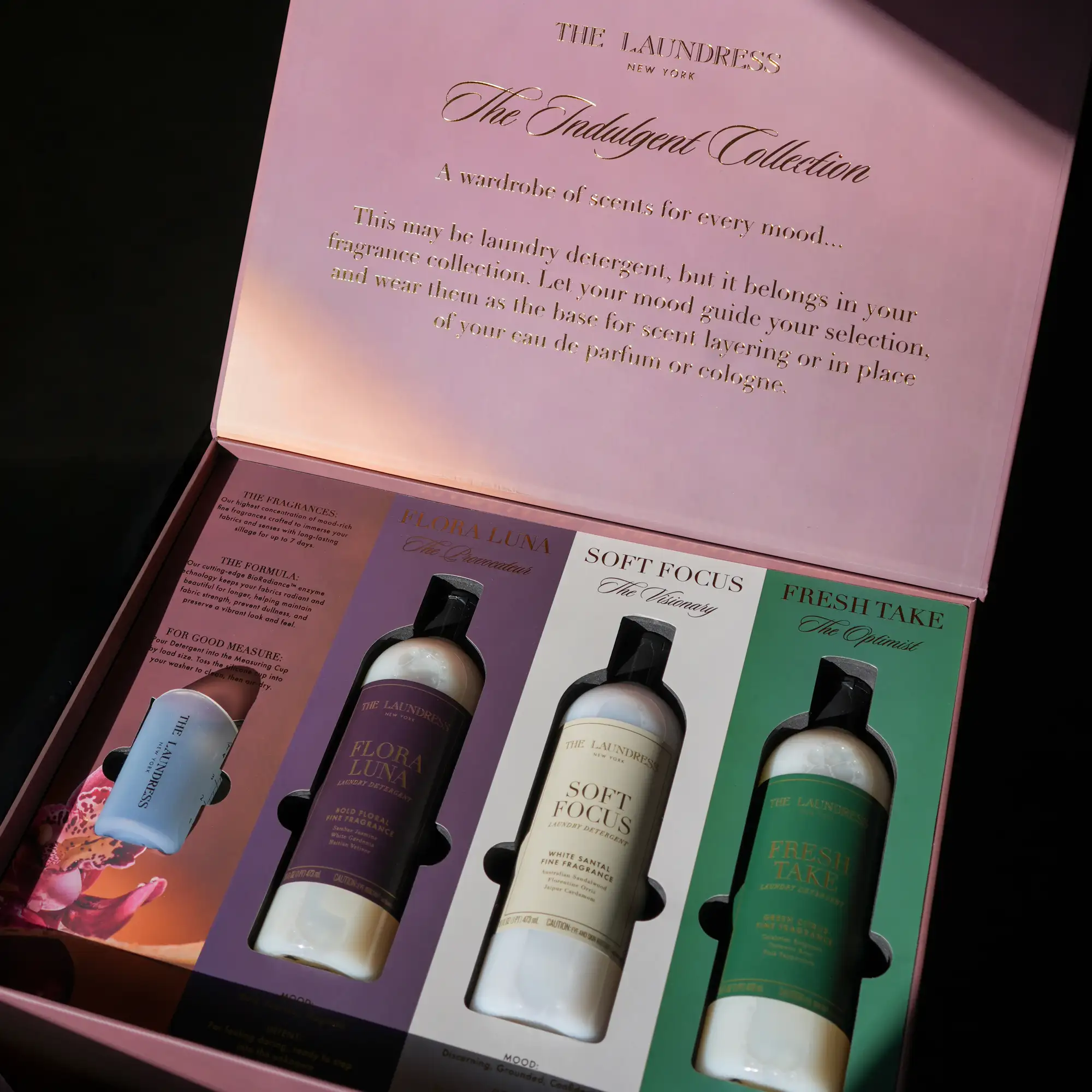 Open pink gift box from The Laundress New York featuring ‘The Indulgent Collection,’ displaying three full-size scented laundry detergents labeled Flora Luna, Soft Focus, and Fresh Take, along with a smaller bottle on the left, all arranged in a colorful, elegant presentation.